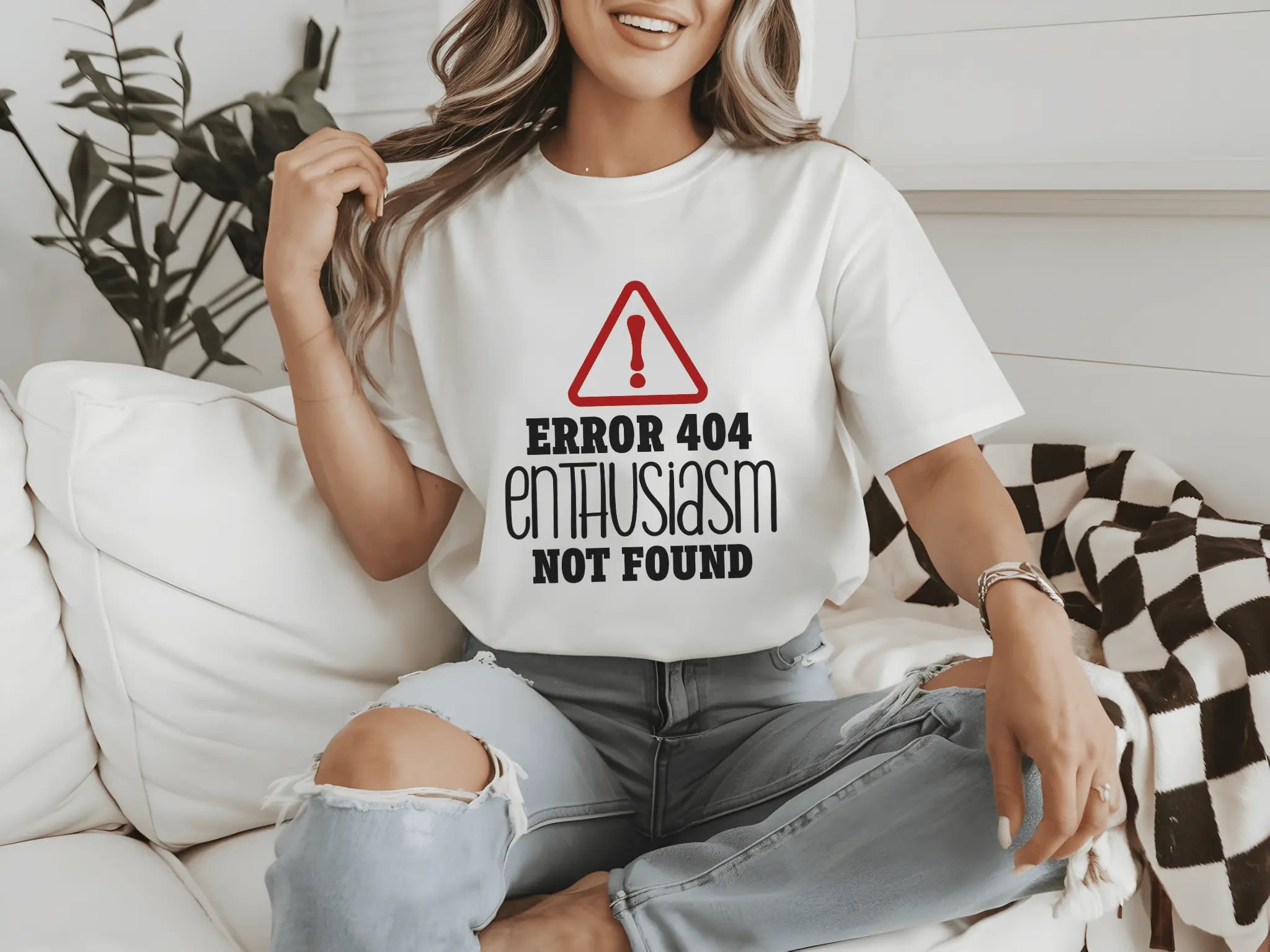 Frau mit weißem „Error 404 Enthusiasm Not Found“-T-Shirt, sitzt auf einem Sofa, trägt zerrissene Jeans und lächelt.
