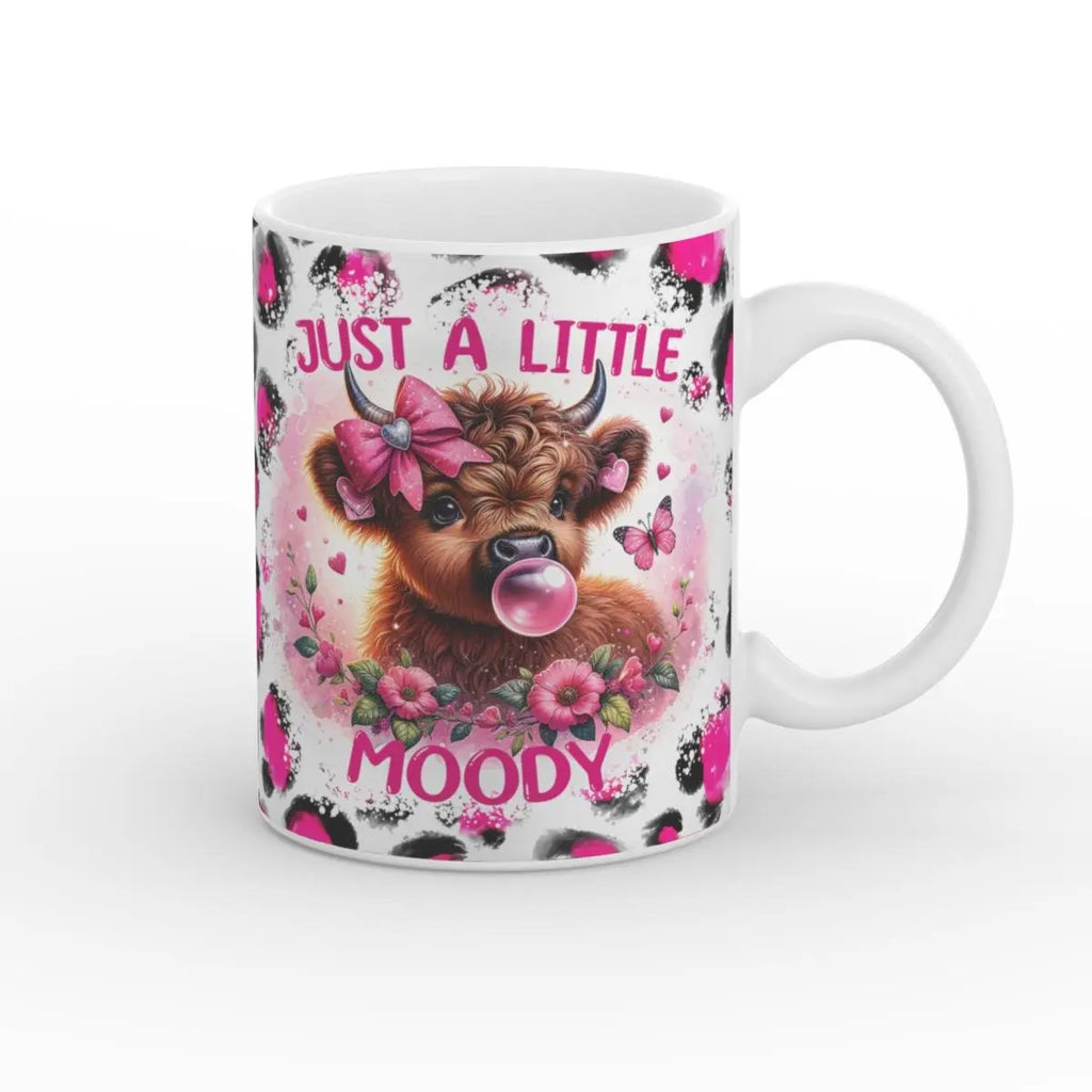 Vorderansicht einer weißen Tasse mit der Highland-Kuh, die eine Kaugummiblase macht, rosa Blumen und der Aufschrift „Just a Little Moody“. Ein charmantes, detailreiches Motiv.